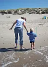 Frau und Kind laufen barfuß am Strand, im Hintergrund Dünen und ein kleines Zelt.