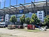 Vor dem Stadion stehen Paletten und Kisten; Arbeiter bereiten den Bereich bei sonnigem Wetter vor.