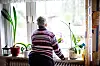 Ältere Dame am Fensterbrett, blickt aus ihrer Wohnung nach draußen, umgeben von Zimmerpflanzen.