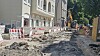 Aufgerissene Straße mit Baustellenabsperrungen, Baggern und Schutt neben Wohngebäuden.