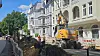 Bagger bei Straßenarbeiten zwischen Häusern, Absperrungen und aufgerissener Fahrbahn.