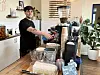 Barista an einer Kaffeemaschine in einem hellen Café mit Tassen und Pflanzen auf der Theke.