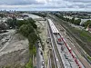 Weitläufiges Bahngelände mit Gleisen, Bahnsteigen und rotem Zug neben Baustellenfahrzeugen.