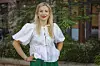 Frau mit weißer Bluse und grünem Rock steht vor Pflanzen und einem Gebäude.