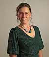 Frau im grünen gepunkteten Kleid mit bunter Halskette vor hellem Hintergrund.