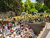 Menschen sitzen und stehen in einem Freiluft-amphitheater, dahinter eine Wiese mit weiteren Besuchern.