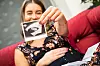 Schwangere Frau auf dem Sofa hält ein Ultraschallbild in die Kamera.
