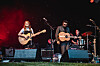 Band spielt mit Gitarren und Schlagzeug auf einer Open-Air-Bühne vor rotem Licht.