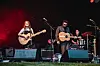 Band spielt mit Gitarren und Schlagzeug auf einer Open-Air-Bühne vor rotem Licht.