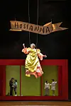 Schwebende Figur unter „Hosianna“-Banner vor gelb-rotem Bühnenbild mit mehreren Personen.