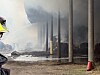 Feuerwehrleute löschen eine brennende Lagerhalle mit Strohballen, dichter Rauch zieht zwischen den Stützen hindurch.