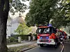 Feuerwehrfahrzeug mit Blaulicht vor einem Haus mit Rauch am Dach, umgeben von Bäumen.