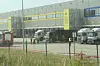Vor einem Gewerbegebäude stehen Lastwagen und Lieferfahrzeuge, einzelne Personen bewegen sich auf dem Vorplatz.
