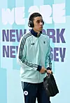 Jamal Musiala in Deutschland-Jacke mit Kopfhörern vor einer „We Are New York New Jersey“-Kulisse.