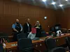 Drei Personen stehen vor Sitzreihen in einem holzgetäfelten Gerichtssaal, eine hält Unterlagen.