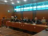 Mehrere Personen sitzen in einem Sitzungssaal an langen Tischen vor großen Fenstern.