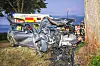 Stark beschädigtes Auto nach einem Unfall neben einem Baum, Einsatzkräfte im Hintergrund.