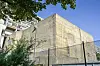 Betonbunker in Schöneberg hinter Zaun und Bäumen unter blauem Himmel.