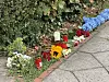 Blumen und brennende Kerzen stehen am Rand eines Gehwegs vor einer Hecke.