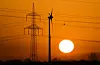 Orangefarbener Sonnenuntergang hinter Strommast, Windrad und Stromleitungen.