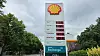 Shell-Tankstelle mit Preistafel an einer Straße, umgeben von Bäumen unter grauem Himmel.