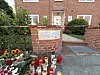 Blumen und Kerzen vor einem brickroten Wohnhaus am Eingang mit Zaun und Mauer.