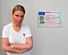 Frau mit verschränkten Armen neben einem EU-Ausweis an grauer Wand.