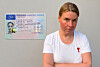 Frau im weißen T-Shirt mit rotem Schleifenabzeichen steht neben einem Ausweis an grauer Wand.