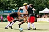 Drei Spieler ringen auf dem Kunstrasen um den Ball, im Hintergrund Werbebanden und ein weißes Zelt.