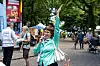 Frau singt mit erhobenem Arm vor Infostand im Park, Besucher gehen über den Weg.