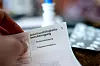 Hand hält ausgefüllte Arbeitsunfähigkeitsbescheinigung, dahinter unscharf Medikamente auf einem Tisch.