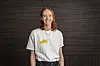 Lächelnde Frau vor dunklem Hintergrund mit weißem T-Shirt und gelbem DBSH-Logo.