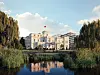 Großes helles Regierungsgebäude mit Flagge, Teich im Vordergrund und Bäumen am Ufer.