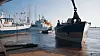Dunkles Schiff am Kai vor mehreren Schiffen und Kränen im Hafen.