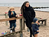 Frau mit zwei Kindern am Holzgeländer am Strand, dahinter Meer und Buhnen.