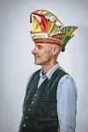 Thomas Hirschbiegel im Profil mit bunter Narrenkappe und Weste vor grauem Hintergrund.