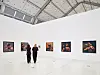 Zwei Besucher gehen durch einen weißen Ausstellungsraum mit mehreren gerahmten Kunstwerken an den Wänden.