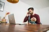 Mann mit Baby telefoniert am Tisch neben offenem Laptop im Wohnraum.