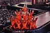 Tanzgruppe in orangefarbenen Kostümen auf einer Bühne vor dichtem Publikum.
