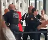 Polizisten und mehrere Personen stehen hinter einer Absperrung in einem Innenraum.