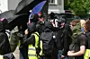 Vermummte Demonstrierende mit schwarzen Schirmen vor einem Gebäude, in der Gruppe ein gelber Warnanzug.