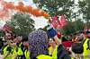 Demonstrierende mit Warnwesten und roten Fahnen vor Baumkulisse, eine Person hält eine Rauchfackel hoch.