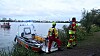 Zwei Einsatzkräfte am Ufer neben einem Rettungsboot, dahinter ein breites Gewässer und Stadtgebäude.