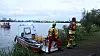 Zwei Einsatzkräfte am Ufer neben einem Rettungsboot, dahinter ein breites Gewässer und Stadtgebäude.