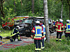Feuerwehrkräfte an einem schwer beschädigten Fahrzeug im Wald, Rettungsgeräte liegen am Boden.