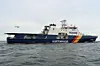 Küstenwachschiff auf ruhiger See, ein Hubschrauber fliegt darüber.