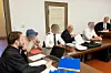 Mehrere Personen beraten an einem langen Tisch mit Laptops, Unterlagen und Sitzungsunterlagen in einem Besprechungsraum.