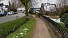 Unbefestigter Weg am Fluss, Häuser links, Brücke und Bäume im winterlichen Ort.