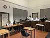 Mehrere Personen im Gerichtssaal an Tischen und im Richterbereich während einer Verhandlung.
