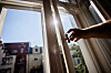 Hand öffnet ein Fenster in heller Wohnung bei sonnigem Wetter; draußen Häuser und blauer Himmel.